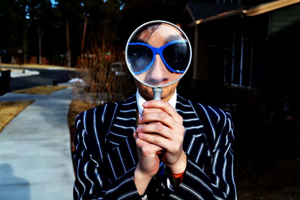 man with shaggy black hair in striped suit holding magnifying glass in front of face enlarging his blue sunglasses