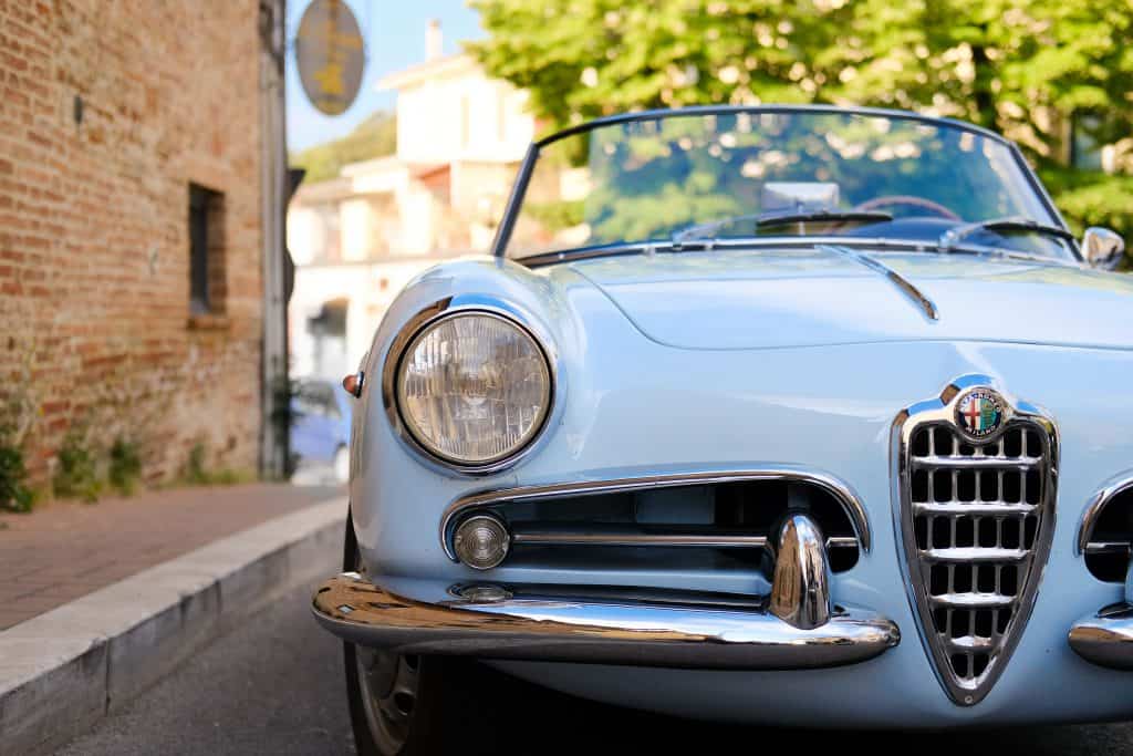 vintage blue convertible with round headlights parked outside next to brick building and tree