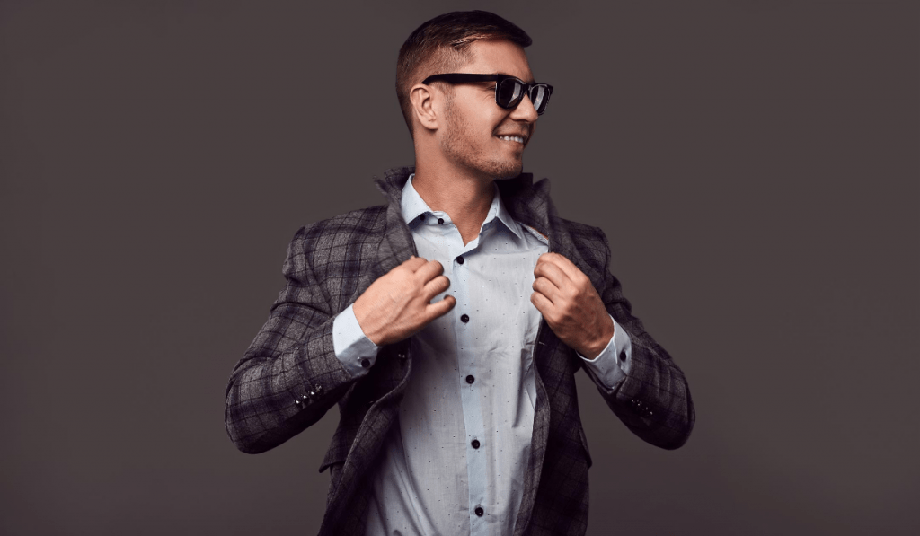 man with thick black glasses wearing a button down shirt and plaid blazer in front of black background