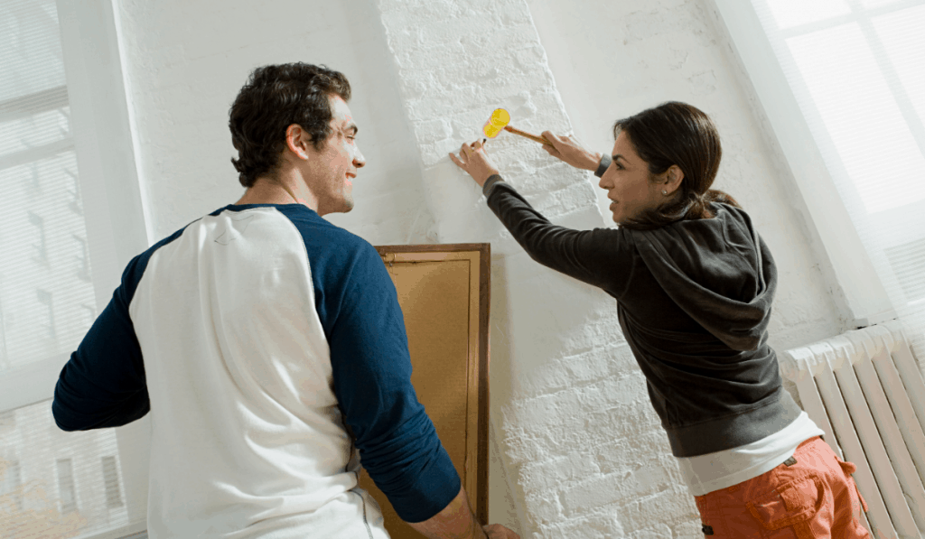 homme femme couple martelant un clou pour accrocher des œuvres d'art sur un mur de briques blanches