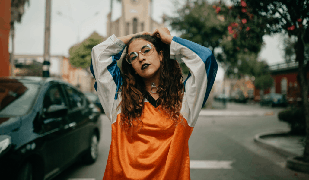 street style girl with round glasses and black lipstick wearing sporty orange and blue nylon pullover standing in street