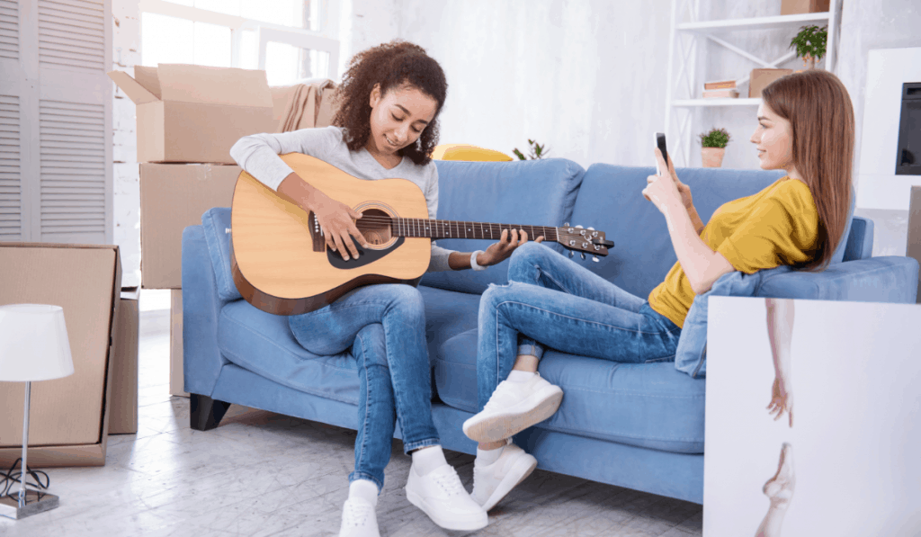 two girls sitting on couch together one playing guitar one looking at phone with moving boxes all around roommates