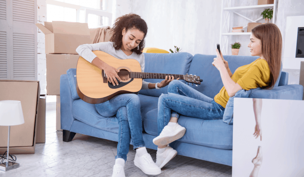 Zwei Mädchen sitzen zusammen auf der Couch, eine spielt Gitarre, die andere schaut auf ihr Telefon und überall stehen Umzugskartons herum