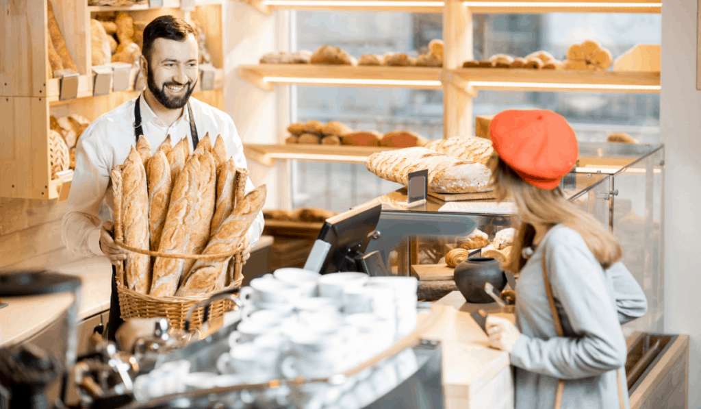 woman in red beret at counter in a bakery talking to man holding a basket of baguettes