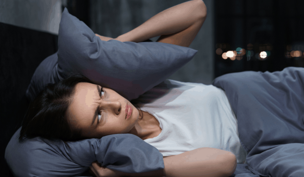 young woman in bed with pillow around head annoyed by neighbors loud music