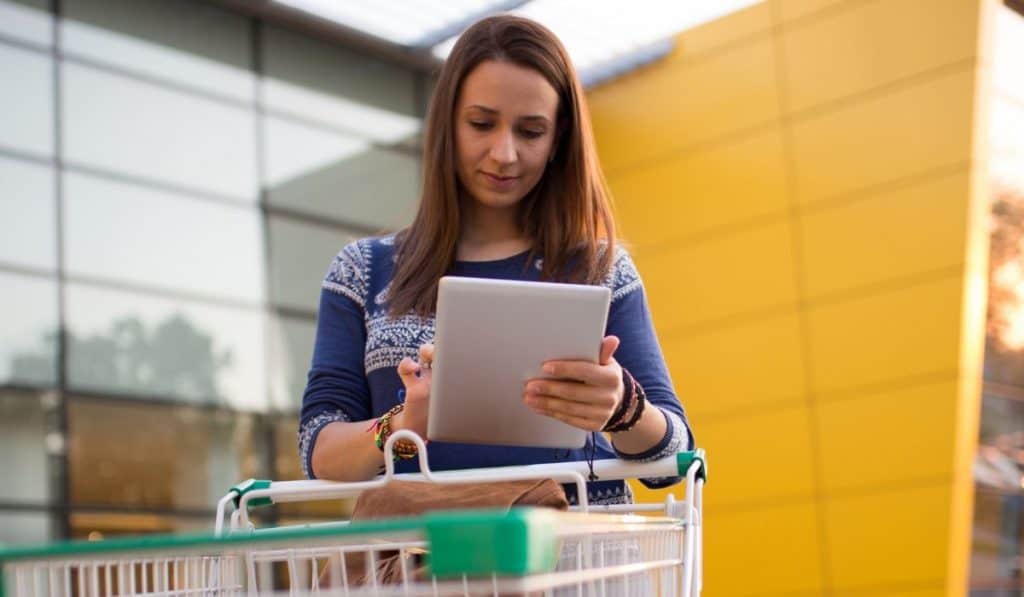 girl outside the mall with a push cart and holding a tablet checking the things she needs for the new place